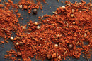 World Spice KC Tailgater's BBQ Rub by World Spice, with its spilled reddish-brown ground spices, whole seeds, and peppercorns scattered across a dark surface, evokes the rich aroma of a classic BBQ rub—perfect for enhancing your favorite dishes.