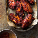 Barbecue chicken wings with a glossy, caramelized coating on parchment paper in a tray, served alongside a small bowl of mixed spices featuring The Spice Lab Ground Star Anise for an inviting, aromatic touch and distinct licorice-like flavor.