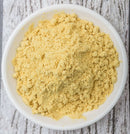 A white bowl filled with The Spice Lab Ground Yellow Mustard 1 lb, a staple among foodservice spices and BBQ seasoning blends from The Spice Lab, sits on a textured, light-colored wooden surface.
