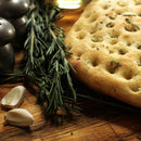 A close-up of fresh focaccia bread topped with The Spice Lab Organic Cut and Sifted Rosemary, garlic cloves, and metal salt and pepper shakers on a wooden surface.