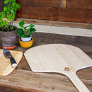 A pair of WPPO LLC Direct 16" x 36" Long Handled Wooden Pizza Peels rests on a rustic wooden counter alongside a small cutting board with a knife, flanked by two potted plants—creating the perfect setup for pizza making.