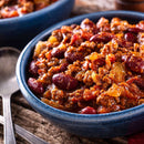 A blue bowl filled with chili made of ground meat, kidney beans, tomatoes, and onions sits on a wooden table with a spoon beside it. The dish is richly flavored with The Spice Lab Organic Chili Powder Blend by The Spice Lab for an authentic Tex-Mex taste.