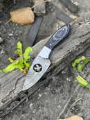 The Texan Knives FIXED BLADE POCKET KNIFE, a personalized Damascus steel hunting knife with a patterned blade and dark handle, lies on weathered wood among green leaves and a dry brown leaf.