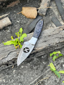 The Texan Knives FIXED BLADE POCKET KNIFE, a personalized Damascus steel hunting knife with a patterned blade and black handle, is displayed outdoors on weathered wood among green leaves and debris.