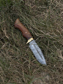 The Texan Knives Hunting Blade is a 10" Damascus steel knife with an engraved blade, rosewood handle, brass guard, and leather sheath, resting on dry, tangled grass.