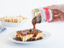 A hand pours Terrapin Ridge Farms Hot Pepper Bacon Jam over cream cheese on a white plate with crackers in the background—an ideal addition to any charcuterie board.