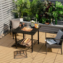A patio scene with the 31 Inch Outdoor Fire Pit Dining Table with Cooking BBQ Grate by DirectDeals, food grilling, drinks, wicker chairs with cushions, firewood stored below, a mesh cover on the deck, and lush potted plants behind.