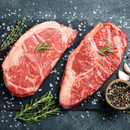 Two raw, marbled Kansas City Strip Steaks from the Dude Food High Steaks Box by Dude Food rest on a slate board, garnished with rosemary sprigs. Coarse salt, garlic cloves, peppercorns, and additional rosemary are scattered around on a textured dark background.