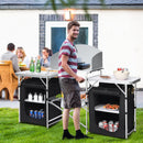 A man grills beside a DirectDeals Folding Camping Table with Storage Organizer holding drinks and supplies, while others enjoy a meal at a backyard table in the grass.