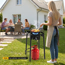 A woman cooks on a DirectDeals Propane Camping Stove 80000 BTU Single Burner with Regulator in her backyard, while four friends chat at a nearby table. A red gas cylinder sits under the stove, with lawn and house in view.
