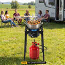 A group sits at a table near an RV on grass. In front, a DirectDeals Propane Camping Stove 80000 BTU Single Burner with Regulator cooks with a pot on the flame.