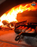 Five sausages sizzle in a cast iron skillet inside a wood-fired oven, with bright flames and burning wood in the background. Enhanced by the WPPO Wood-Fired Oven Flame / Heat Deflector from WPPO LLC Direct—a must-have pizza oven accessory—this setup ensures even cooking and perfect results. The WPPO pizza oven logo appears in the upper right corner.