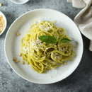 A plate of spaghetti with green pesto sauce, garnished with grated cheese, The Spice Lab Organic Pine Nuts by The Spice Lab, and a fresh basil leaf, on a white plate against a gray background. The Spice Lab Organic Pine Nuts add both flavor and texture to the dish, making it perfect as a hearty meal or an inspiring salad topping.