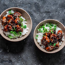 Two bowls of rice topped with glazed chicken, seasoned with a hint of The Spice Lab Organic Ground Ginger and sprinkled with sesame seeds, are garnished with fresh greens and set on a dark textured surface.