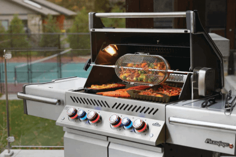 A stainless steel outdoor grill with four control knobs is cooking assorted vegetables in a rotisserie basket and trays of food on the grill grates, set on a patio with a pool in the background.