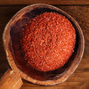 A wooden scoop filled with The Spice Lab Hawaiian Red Alaea Sea Salt Fine Grain sits on a wooden surface.