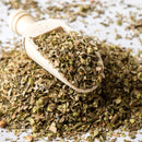 A wooden scoop brimming with The Spice Lab Organic Oregano Leaf rests atop a mound of the same fragrant herb, with loose leaves and seeds from The Spice Lab scattered over a white surface—a classic staple in Italian cooking.