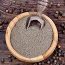 A rustic wooden bowl filled with The Spice Lab Organic Ground Black Pepper and a small wooden scoop, surrounded by whole organic peppercorns on a weathered wooden surface.