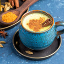 A blue mug filled with golden milk topped with cinnamon and The Spice Lab Organic Cinnamon Stick, placed on a saucer with cardamom pods nearby. Turmeric powder, chai spices, and The Spice Lab cinnamon are in the background.