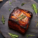 A piece of roasted meat with a charred crust sits on a dark round board, garnished with sprigs of fresh rosemary and thyme, and surrounded by extra herbs and grilling spices from The Spice Lab Best Seller Seasoning Set by The Spice Lab.
