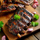 Grilled barbecue pork ribs garnished with fresh parsley on a wooden cutting board, with The Spice Lab Organic Minced Garlic and parsley leaves scattered around.