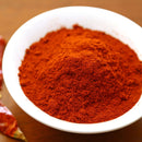 A white bowl filled with vibrant red The Spice Lab Cayenne Pepper X-Hot Ground sits on a wooden surface, with dried red chili peppers visible beside it.