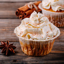 A cupcake topped with swirled whipped cream and a sprinkle of The Spice Lab Apple Pie Spice Blend sits on a wooden surface, with cinnamon sticks and a star anise in the background.
