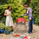 A woman in a white dress and a man in a blue shirt cook on the DirectDeals 2-in-1 Gas Camping Grill and Stove with, surrounded by flowers, dishes, and fresh vegetables. She holds a drink while he tends the grill.