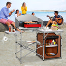 The DirectDeals Foldable Outdoor BBQ Portable Grilling Table With Windscreen Bag is set up on a beach, neatly holding food and supplies as four people enjoy music and laughter in the background.
