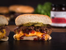 A cheeseburger sliced in half reveals melted cheese, pickles, sauce, and a layer of Terrapin Ridge Farms Hot Pepper Bacon Jam inside the beef patty on a bun, with a blurred jar of Terrapin Ridge Farms and greenery in the background.