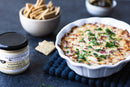 A white baking dish of savory dip, topped with herbs, sits on a blue mat beside crackers, pickles, and a jar of Terrapin Ridge Farms Roasted Garlic Horseradish Dip—perfect as a sandwich spread.