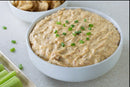A white bowl of Terrapin Ridge Farms Hatch Chile Cream Cheese Dip, topped with chopped green onions, sits next to a bowl of pork rinds and a tray of celery sticks on a light countertop.