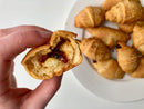 A hand holds a partially eaten crescent roll filled with jam and cheese, featuring Terrapin Ridge Farms Hot Pepper Bacon Jam - 5 oz for a sweet and savory flavor. In the background, whole crescent rolls rest on a plate atop a white surface.