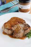 A grilled pork chop topped with sweet and savory Terrapin Ridge Farms Pecan Honey Mustard is served on a white plate with parsley. A jar of Terrapin Ridge Pecan Honey Mustard appears in the background.