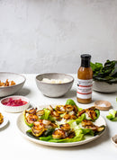 Grilled shrimp skewers on lettuce rest on a white table, with bowls of rice, greens, pickled onions, and a bottle of Terrapin Ridge Farms Tropical Tequila Sauce by Terrapin Ridge Farms. A light-colored wall forms the background.