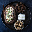 A round tray holds a bread loaf with savory dip, a bowl of crackers, dark bread cubes, and a jar of Terrapin Ridge Farms Roasted Garlic Horseradish Dip by Terrapin Ridge Farms on a dark surface.