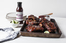 A tray of glazed ribs with lime wedges sits on a white table, next to Terrapin Ridge Farms Raspberry Peach Chipotle Sauce—a perfect barbecue glaze—plus limes, plates, and a striped cloth. A brush rests on the tray.