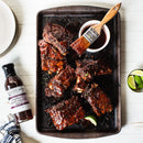 A tray of glazed barbecue ribs with a brush in a cup of Terrapin Ridge Farms Raspberry Peach Chipotle Sauce, plus lime wedges and a bottle of Terrapin Ridge Farms sauce on the side, showcase this bold glaze on a white wooden surface.