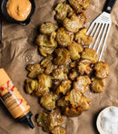 Crispy smashed potatoes sprinkled with salt are arranged on parchment paper with a spatula, Terrapin Ridge Farms Sriracha Horseradish Garnishing Squeeze Sauce for drizzling, a bowl of creamy dip, and a dish of flaky salt.