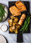 Two cooked salmon fillets on a dark cutting board with roasted smashed potatoes, sautéed green beans, and a bottle of Terrapin Ridge Farms Sriracha Horseradish Garnishing Squeeze Sauce—all arranged on a marble surface.