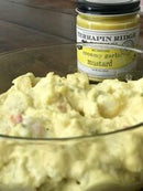 A glass bowl of creamy potato salad sits in the foreground, with a jar of Terrapin Ridge Farms Creamy Garlic Mustard blurred in the background on a wooden surface.