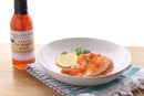 A plate with chicken breast, orange sauce, lemon, and cilantro sits beside a bottle of Terrapin Ridge Farms Hot Pepper Peach Bourbon Sauce on a striped towel.