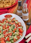 A white bowl of salad sits on a colorful striped tablecloth beside two bottles of Terrapin Ridge Farms Garlic & Herb Vinaigrette and a platter of pita chips in the background.