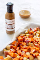 A bottle of Terrapin Ridge Farms Apple Cider Maple Balsamic Vinaigrette stands beside a glass container and a baking sheet filled with colorful roasted potatoes and sweet potatoes.