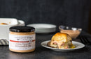 A jar of Terrapin Ridge Farms Onion Blossom Horseradish Dip sits beside a slider sandwich with meat, cheese, and sauce, all set on a dark surface with blurred plates and dipping sauces in the background.