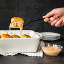 A hand lifts a cheesy slider from a white baking dish, with Terrapin Ridge Farms Onion Blossom Horseradish Dip in a glass bowl and a stack of white plates nearby on a striped cloth.