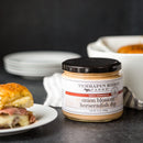 A jar of Terrapin Ridge Farms Onion Blossom Horseradish Dip sits beside a sandwich, with plates and a baking dish of bread rolls in the background.