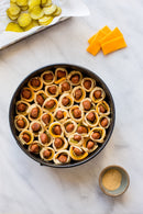 A round baking pan of unbaked pigs in a blanket with cheese sits near pickle slices and extra cheese, alongside a bowl of Terrapin Ridge Farms Raspberry Honey Mustard Pretzel Dip for a sweet and tangy flavor.