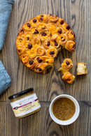A round pull-apart cheese and sausage bread sits on wood with pieces pulled out. Next to it is a jar of Terrapin Ridge Farms Raspberry Honey Mustard Pretzel Dip and a bowl of coarse mustard, with a blue cloth nearby.