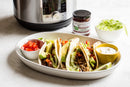 A plate holds four tacos filled with meat, avocado, and veggies, served with bowls of sour cream and diced tomatoes. Nearby: shredded lettuce, salsa, and a slow cooker with Terrapin Ridge Farms Jalapeno Hatch Chile Jam for sweet heat.